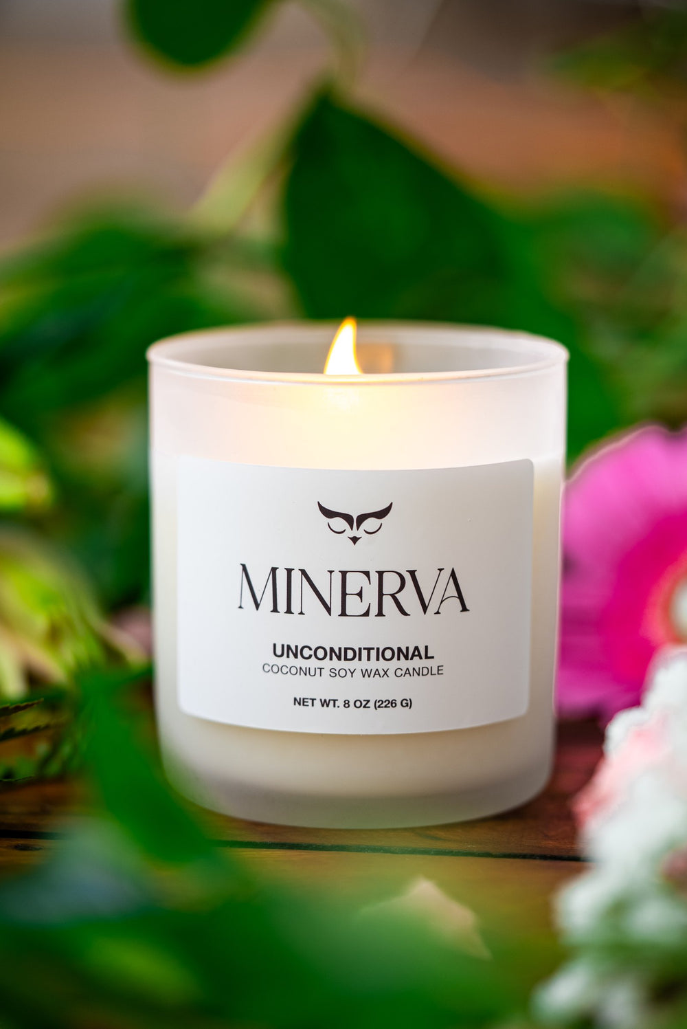 Lit Minerva Unconditional candle among pink flowers