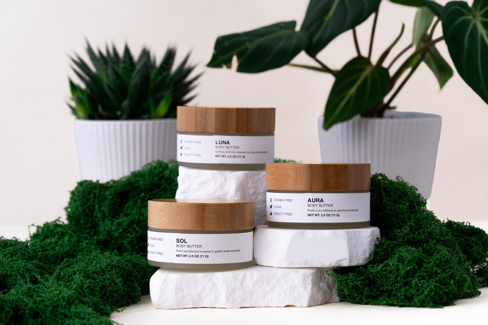Three Minerva body butter jars with bamboo lids arranged on white stone and green moss with plants