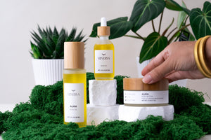 Hand reaching for a Minerva body butter jar beside Aura body oil on green moss