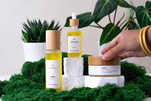 Hand reaching for a Minerva body butter jar beside Aura body oil on green moss