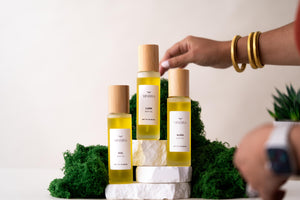 Hand reaching for one of three Minerva botanical body oils on white stone and green moss