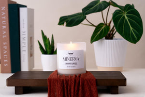 Lit Minerva Harvest Moon candle on a wooden tray with a rust linen cloth, books and plants