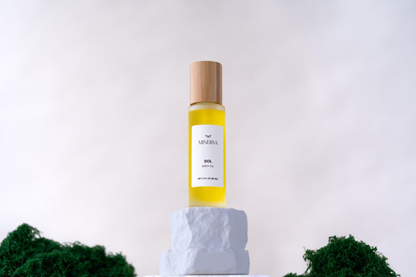 Minerva Sol body oil in a frosted glass bottle on a white plinth with moss