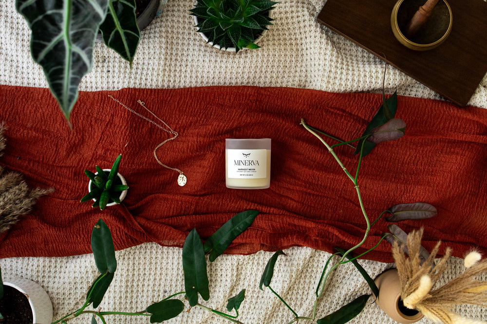 Lit Minerva Harvest Moon candle on a wooden tray with a rust linen cloth, books and plants